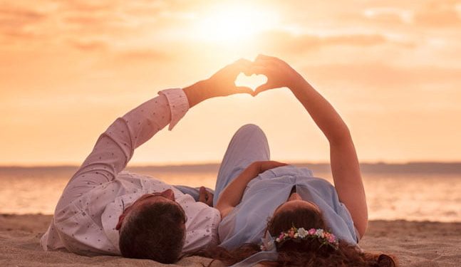 couple laying on beach at sunset making a heart with their hands