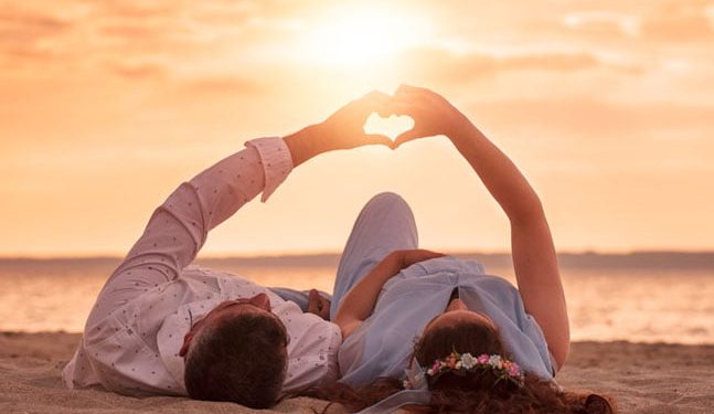 couple-laying-on-sand-by-water-5cc868f41a499 couple laying on beach at sunset making a heart with their hands