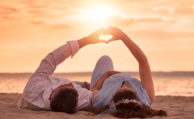 couple laying on beach at sunset making a heart with their hands