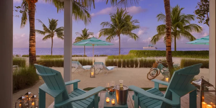 A patio with lounge chairs and champagne with a view of the beach, palm trees, umbrellas, and the ocean.