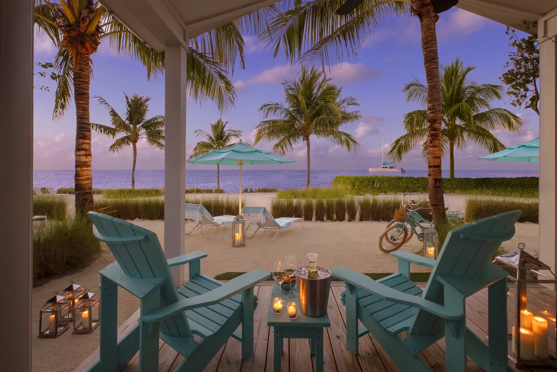 A patio with lounge chairs and champagne with a view of the beach, palm trees, umbrellas, and the ocean.